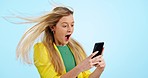 Surprise, wind and woman with phone in studio for wow, news or social media promo on blue background. Mind blowing, omg or female model with smartphone, app and excited by sale, deal or sign up offer