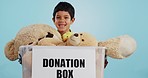 Boy in studio with toys, donation in box and smile for children, social charity and hope in kindness. Care, donate and happy kindergarten child with teddy bear package at kids ngo on blue background.