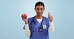 Face, woman and doctor with thumbs up, apple and eating with wellness on a blue studio background. Person, model or medical professional with a fruit, nutrition or delicious with food or healthy diet