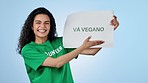 Volunteer woman, vegan board and studio for smile on face, cheers and pointing by blue background. Recruitment, diet choice and call to action for sustainable food, fist sign or poster in Barcelona