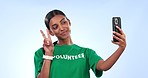 Volunteer woman, selfie and smile in studio with peace sign, emoji and pride for community service by blue background. Girl, NGO worker and happy with icon, review and v for winning on social network