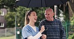 Charity, umbrella and a nurse walking with an old man outdoor in the garden or park for senior care. Smile, conversation or assistance with a senior patient and volunteer outdoor for wellness