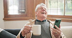 Coffee, phone and funny senior man on sofa, streaming comedy movie or video in living room at home. Drink tea, smartphone and person laugh on couch for social media meme, reading email or joke on app