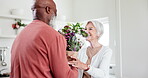 Senior, couple and flowers for love, care or happiness with surprise, gift or birthday in kitchen of home. Elderly, black man and woman or interracial with bouquet for celebration, present or date