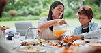Family, pouring and orange juice at table for lunch, party or event with eating, bonding and conversation. People, food and beverage in jug and glass for dinner, nutrition and meal outdoor in garden