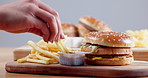 Person, hand and fast food with chips, burger and sauce for delicious meal, snack or lunch against a gray studio background. Closeup of customer dipping french fries for eating, advertising or mockup