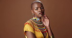 African fashion, face and black woman in studio with stylish, beautiful and traditional outfit. Serious, pride and portrait of young female model with trendy clothes for culture by brown background.
