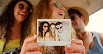 A cheerful young woman holding a polaroid of herself and her friends sitting in a camper van