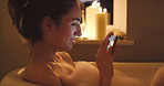 A young woman scrolling on her mobile phone during a nighttime bubble bath