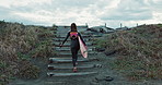 Back, woman and surfboard at beach, walking up stairs and ready for fitness with wet suit, sports and surfing in nature. Steps, exercise and surfer, ocean with professional or hobby with challenge