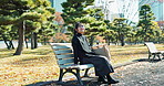 Japanese woman, bench and smartphone in park with relax, happiness and social media in sunshine in city. Female person, sitting and wellness with cellphone in town, streaming and digital technology