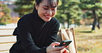 Happy asian woman, phone and sitting on park bench for social media, communication or networking. Female person smile in relax on mobile smartphone for online search, streaming or reading in nature