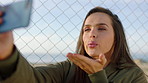 A happy young woman waving and blowing kisses during a video call on her smartphone. A young woman standing by a fence making a video call on her cellphone and blowing a kiss and waving.