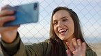 A happy young woman making a video call on her cellphone and waving. A woman waving during a video call made on her smartphone and standing by a fence. A smiling woman talking during a video call