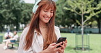 Woman, cellphone and headphones or texting in park or smile or online communication, streaming or playlist. Female person, smartphone and message in nature or relax weekend or podcast, track or song
