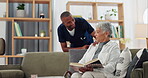 Senior, woman and caregiver on sofa with support, conversation and nurse in living room of retirement home. Elderly, person and black man with kindness, happiness and discussion while reading a book