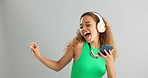 Happy woman, phone and listening with headphones to music and dancing on a gray studio background. Excited female person or model enjoying audio podcast or sound on mobile smartphone or mockup space