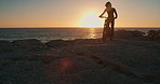 Person, hill and motorbike with sunset for travel, adventure or riding on rocks, training or extreme sports. Biker silhouette with safety helmet and bike for journey by ocean or sea in lens flare