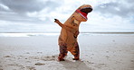 Animal, funny and dinosaur costume on beach dancing with energy for comedy joke on vacation travel. Goofy, silly and inflatable reptile mascot moving, jumping and having fun by ocean on holiday.
