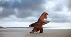 Surfboard, outdoor and dinosaur costume on beach for water sports competition training for comedy. Goofy, running and inflatable t rex mascot by ocean or sea for surfing with comic or funny joke.