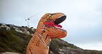 Funny, costume and inflatable dinosaur outdoor at beach with mountain and nature in background. Comedy, character and monster with teeth in jaws and morning with blue sky on holiday or vacation