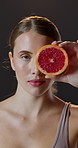 Woman, face and skincare with grapefruit in vertical portrait for fruit detox and natural treatment in studio. Female person, beauty and healthy skin from vitamin A and antioxidant on dark background