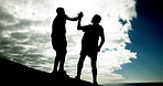 People, high five and fitness on rock at beach for helping hand, clouds and outdoor together. Men, support and lift for partnership, silhouette or low angle in training, exercise or workout in summer