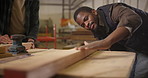 Black man, carpenter and blowing dust with wood at workshop in crafting, precision or sanding. Young African, artisan or woodworker checking plank, board or flat log on workbench at storage warehouse