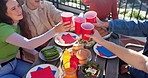 Cheers, alcohol and friends with food outdoor for hamburger lunch, party or gathering together. Happy, toast and group of young people with drinks and meal on balcony for bonding by wooden table.