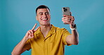 Man, peace sign and phone for selfie in studio, live streaming and emoji on blue background. Male person, symbol and photography for profile picture, social media update and capture memory online