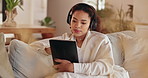 Home, headphones and woman on sofa, tablet and typing with sound, live streaming and listening to music. Apartment, person and girl on couch, tech and headset for podcast, rest and comfort in lounge