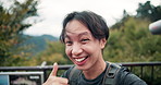 Happy, asian man and nature with thumbs up for video call, travel or hiking adventure in forest. Portrait, pov or japanese male person with smile for live recording, trip or exploration in woods