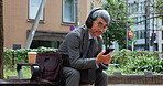 Mature man, headphones and music on business trip in city with phone, travel or reading on sidewalk bench. Professional person, chat and waiting for transport with sound, streaming or road in Japan