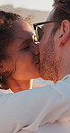 Love, couple and happy with kissing by beach for romantic date, anniversary travel and bonding together. Breeze, hug and interracial people with affection in nature for holiday, loyalty or commitment