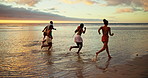 Beach, sunset and friends running on sea water for adventure, freedom and celebration with energy. People, ocean race and group on trip outdoor with back view for summer vacation, travel and holiday