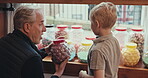 Grandfather, shopping and choice of candy for a child at a shop or sweets store shelf. Senior man and a boy kid or customer talking and buying sugar treats in window display jar on travel trip