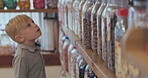 Kid, shopping in a candy store and choice of sweets, confectionery or boy in a retail supermarket, shop and sale of candies. Jar, shelf and child choosing product, chocolate or toddler with decision