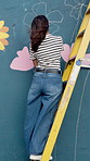 Artist, color and painting on mural with ladder for flower decor, back and volunteer with creative design in city. Art, woman and painter with brush on wall for community project, charity and wind