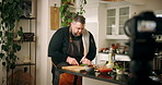 Man, chef and recording in kitchen with cutting, board and herbs for flavor with meal prep on cooking show. Person, knife and cuisine with produce, ingredients and recipe for live streaming in home