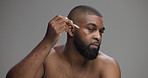 Face, skincare and man with beauty serum for collagen treatment or dermatology. African person, dropper and apply facial cosmetics for hydration or glow with hyaluronic acid on gray studio background