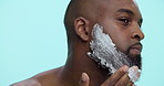 Black man, beard or grooming with shaving cream for hair removal on a blue studio background. Male person, model and applying foam or aftershave with soap for epilation, facial cleaning or hygiene