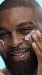 Black man, hands and lotion on face in studio for skincare, cosmetics or moisturizer on blue background. Smile, portrait and male person apply cream, sunscreen or facial treatment for dermatology