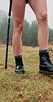 Person, legs and hiker with walking stick on grass field for adventure, journey or outdoor trip. Closeup, hiking or trekking with boots, shoes or aid for physical activity or exercise in forest