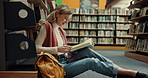 Happy woman, student and reading with book in library on floor for education, learning or knowledge. Female person, learner or sitting with novel or textbook for literature or studying by bookshelf