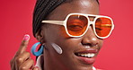 Cream, happy and face of black woman for sunscreen, beauty and facial treatment in studio. Sunglasses, apply and portrait of person with moisturizer, lotion and smile for skincare on red background