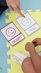 Hands, child therapist and card with emotion for decision, guide and test for wellness at clinic. Psychologist, kid and above with happy, sad or assessment with emoji for mental health exam on floor