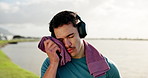 Fitness, music and towel with man outdoor for break from cardio challenge or intense training. Headphones, sweating and thinking with tired athlete at lake for exercise or recovery from workout