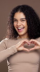 Face, heart hands and love with happy woman in studio for kindness or excited on brown background. Portrait, smile and girl with romance sign for care, support or laugh with dance for thank you emoji