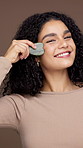 Face, gua sha and smile of woman in studio to relax skin for beauty, cosmetics or dermatology. Massage, skincare and stone tool with happy model isolated on brown background for facial treatment