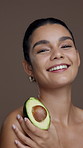 Avocado, beauty and smile with face of woman in studio for natural skincare, self care and cosmetics. Glow, omega 3 benefits and vitamin e with person and fruit on brown background for antioxidants